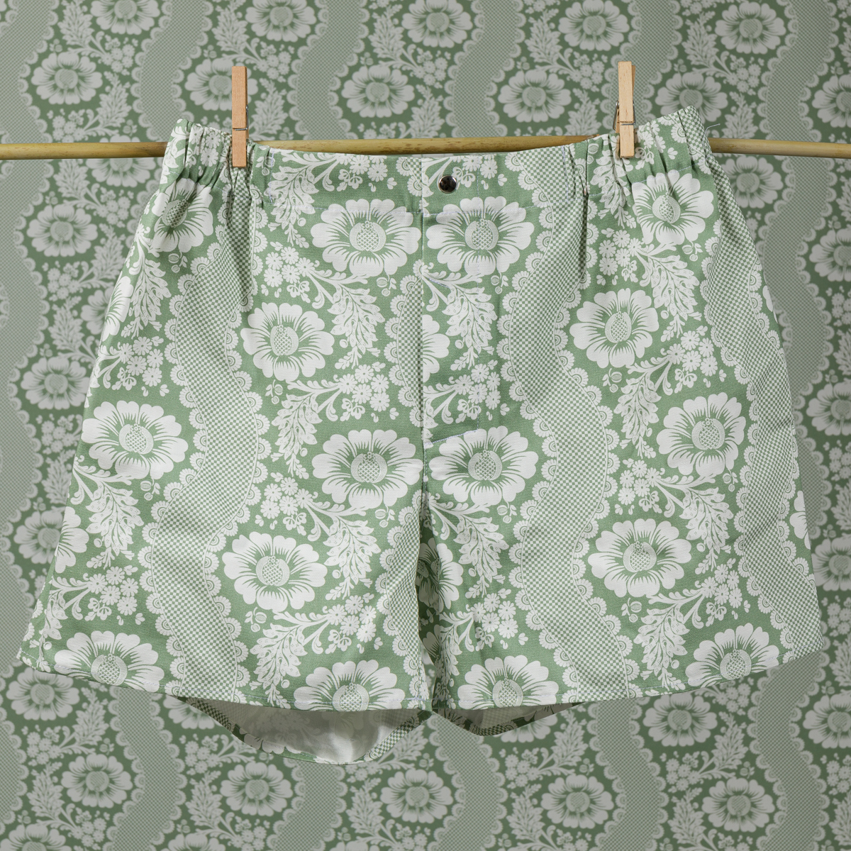 Le short CALEçON « FLEURS STYLISÉES » SAUGE, vert et blanc, est suspendu à une corde à linge devant un mur recouvert d'un papier peint à motifs floraux assortis, les deux éléments se fondant l'un dans l'autre pour créer un effet de camouflage. la marque Toile de Jouy