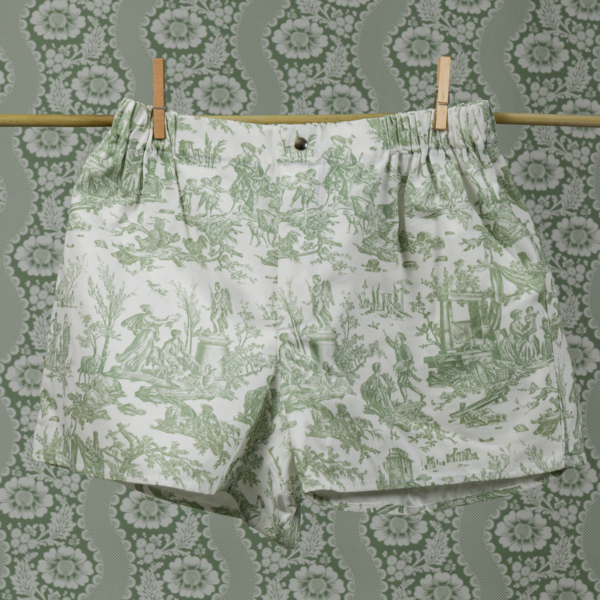Le boxer CALEçON « FLEURS STYLISÉES » SAUGE, orné de motifs de toile de Jouy verts évoquant la campagne, est suspendu à une corde à linge à l'aide de pinces à linge. L'arrière-plan présente des motifs floraux et paysagers verts et blancs assortis qui s'harmonisent avec le boxer. la marque Toile de Jouy