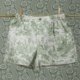 Le boxer CALEçON « FLEURS STYLISÉES » SAUGE, orné de motifs de toile de Jouy verts évoquant la campagne, est suspendu à une corde à linge à l'aide de pinces à linge. L'arrière-plan présente des motifs floraux et paysagers verts et blancs assortis qui s'harmonisent avec le boxer. la marque Toile de Jouy