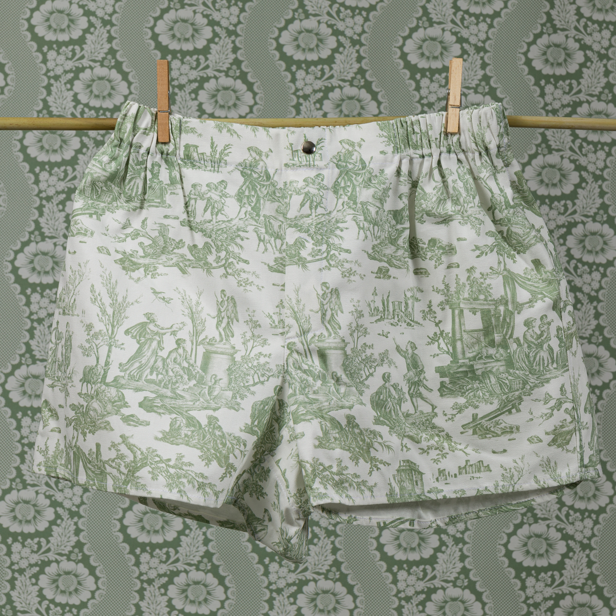 Le boxer CALEçON « FLEURS STYLISÉES » SAUGE, orné de motifs de toile de Jouy verts évoquant la campagne, est suspendu à une corde à linge à l'aide de pinces à linge. L'arrière-plan présente des motifs floraux et paysagers verts et blancs assortis qui s'harmonisent avec le boxer. la marque Toile de Jouy