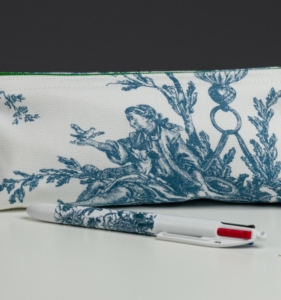 Le XCOMM_BIC ESCARPOLETTE BLEU, un stylo blanc orné d'un motif bleu, repose sur une surface blanche devant son étui assorti, décoré d'une illustration bleue représentant une personne assise tendant la main vers un oiseau. la marque Toile de Jouy