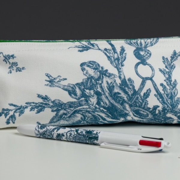 Le XCOMM_BIC ESCARPOLETTE BLEU, un stylo blanc orné d'un motif bleu, repose sur une surface blanche devant son étui assorti, décoré d'une illustration bleue représentant une personne assise tendant la main vers un oiseau. la marque Toile de Jouy