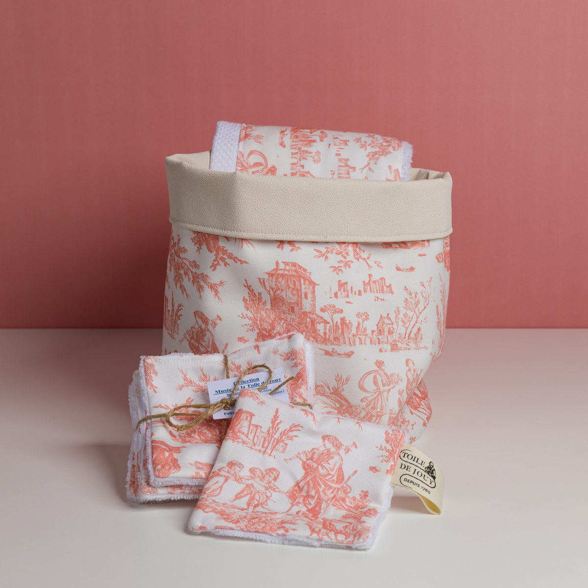 Le panier de rangement en tissu XCOMM_VANITY OFFRANDE SAUGE, ainsi que les linges pliés, attachés avec de la ficelle ou étiquetés, arborent un motif de toile de Jolis champêtres rose et blanc sur un fond blanc avec une base rose. la marque Toile de Jouy