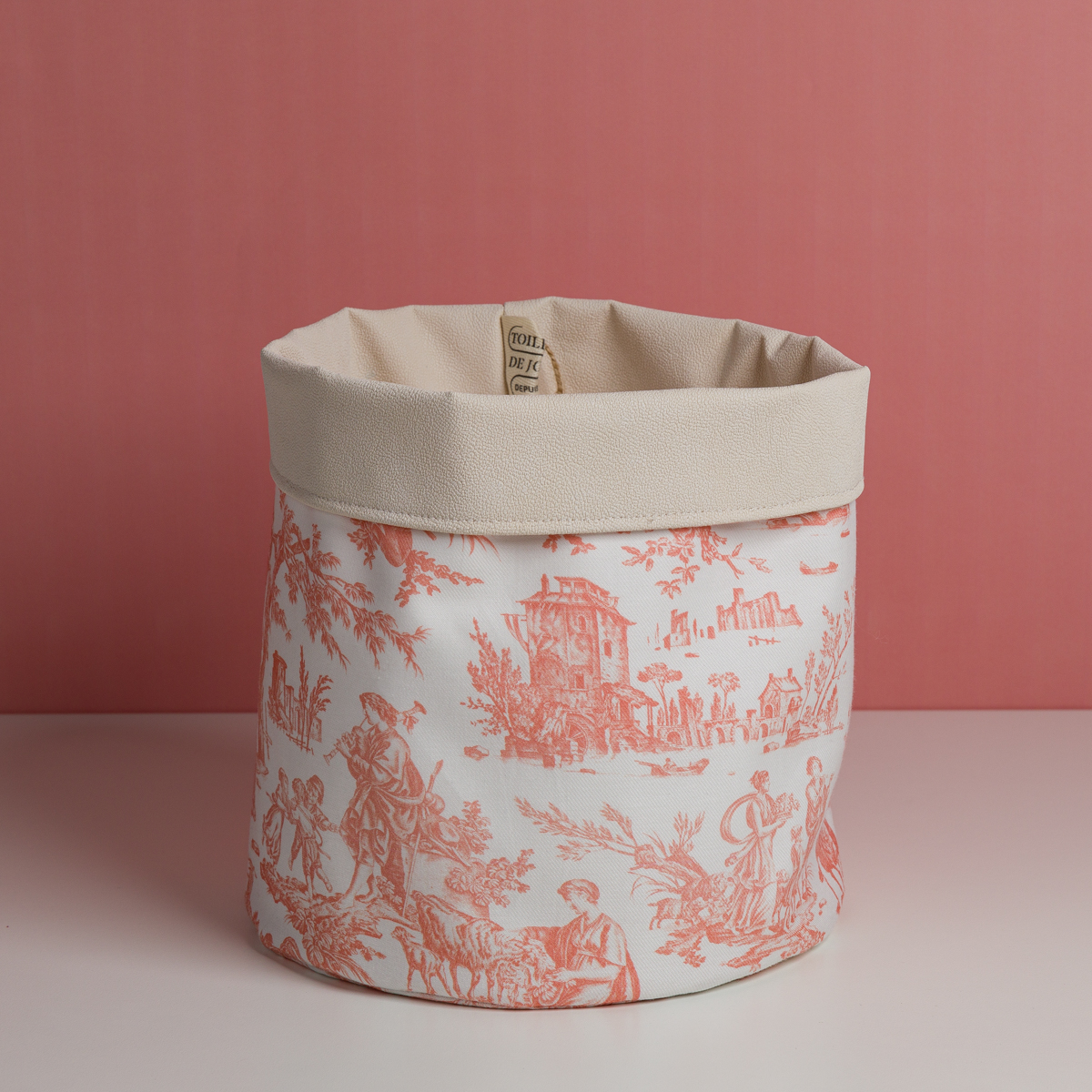 Le modèle XCOMM_VANITY OFFRANDE SAUGE est un panier de rangement décoratif en tissu, doté d'un bord supérieur de couleur crème et d'un motif « toile pastorale » rouge sur fond blanc, présenté sur un fond rose. la marque Toile de Jouy