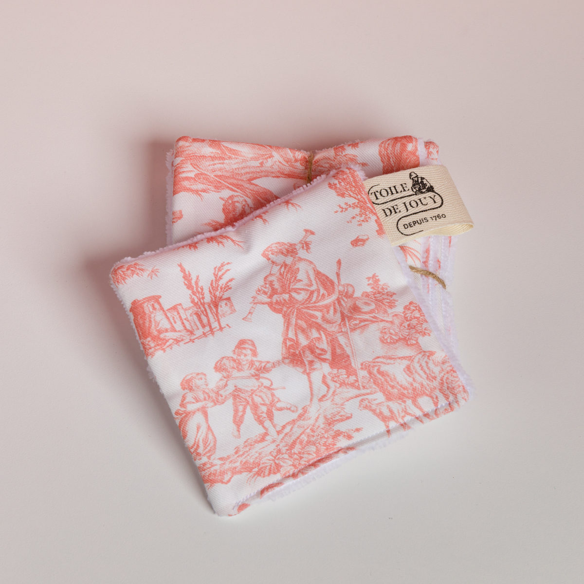 Deux carrés de tissu pliés de la collection XCOMM_VANITY OFFRANDE SAUGE arborent un imprimé « toile de Jouy » bucolique rouge et blanc, orné de personnages champêtres. L'un des carrés porte une étiquette beige sur laquelle on peut lire « TOILE DE JOUY DEPUIS 1760 ». la marque Toile de Jouy