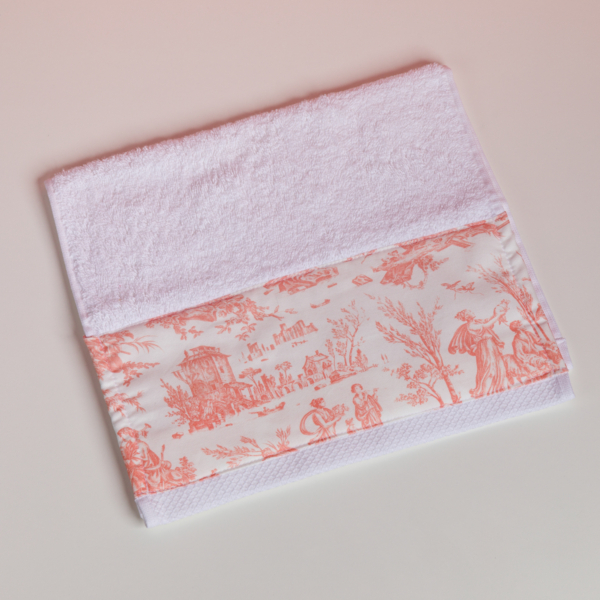 Le modèle XCOMM_VANITY OFFRANDE SAUGE est une serviette blanche ornée d'une bordure représentant une scène champêtre en rose et blanc, avec des personnages, des arbres et des bâtiments, posée à plat sur une surface claire. la marque Toile de Jouy