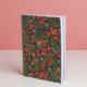 L'EMPREINTE SOUS PASSE-PARTOUT "BONNES HERBES - BLEUETS" est un carnet de notes sur pied avec une couverture florale vibrante dans les tons rouge, jaune et bleu. Il contient du papier millimétré et se présente sur un fond rose. la marque Toile de Jouy