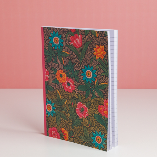 Le carnet EMPREINTE SOUS PASSE-PARTOUT "BONNES HERBES - BLEUETS" présente une couverture florale vibrante avec des fleurs rouges, orange et bleues. Il se tient debout sur une surface blanche devant un fond rose, et contient des pages quadrillées. la marque Toile de Jouy