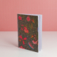 Le carnet à couverture rigide EMPREINTE SOUS PASSE-PARTOUT "BONNES HERBES - BLEUETS", au motif floral coloré, se tient debout sur une surface blanche sur fond rose, ses pages lignées étant partiellement ouvertes. la marque Toile de Jouy