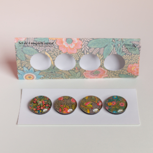 L'EMPREINTE SOUS PASSE-PARTOUT "BONNES HERBES - BLEUETS" est un ensemble de quatre aimants ronds en cristal avec des motifs floraux, présentés sur une carte blanche devant un emballage à motifs floraux avec quatre découpes circulaires. la marque Toile de Jouy