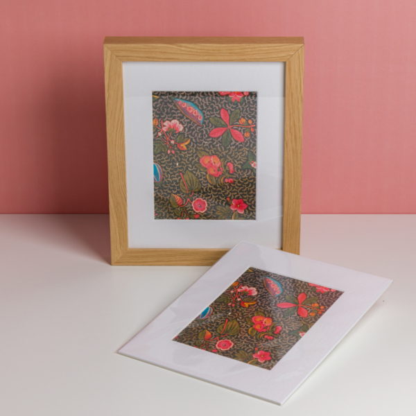 L'EMPREINTE SOUS PASSE-PARTOUT "BONNES HERBES - BLEUETS" présente une œuvre d'art floral encadrée avec des fleurs rouges, roses et bleues sur un fond sombre, exposée avec un imprimé assorti à côté sur un mur rose. la marque Toile de Jouy