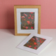 L'EMPREINTE SOUS PASSE-PARTOUT "BONNES HERBES - BLEUETS" présente une œuvre d'art floral encadrée avec des fleurs rouges, roses et bleues sur un fond sombre, exposée avec un imprimé assorti à côté sur un mur rose. la marque Toile de Jouy