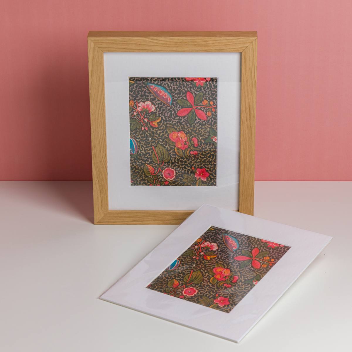 L'EMPREINTE SOUS PASSE-PARTOUT "BONNES HERBES - BLEUETS" présente une œuvre d'art floral encadrée avec des fleurs rouges, roses et bleues sur un fond sombre, exposée avec un imprimé assorti à côté sur un mur rose. la marque Toile de Jouy