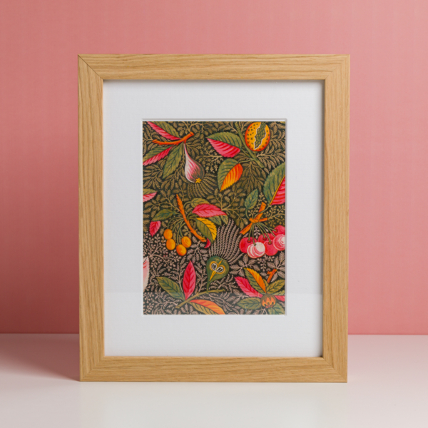 EMPREINTE SOUS PASSE-PARTOUT 'BONNES HERBES - BLEUETS' est une œuvre d'art encadrée avec une bordure en bois présentant une illustration botanique vibrante de feuilles, de baies et de fleurs colorées sur un fond sombre contre un mur rose. la marque Toile de Jouy