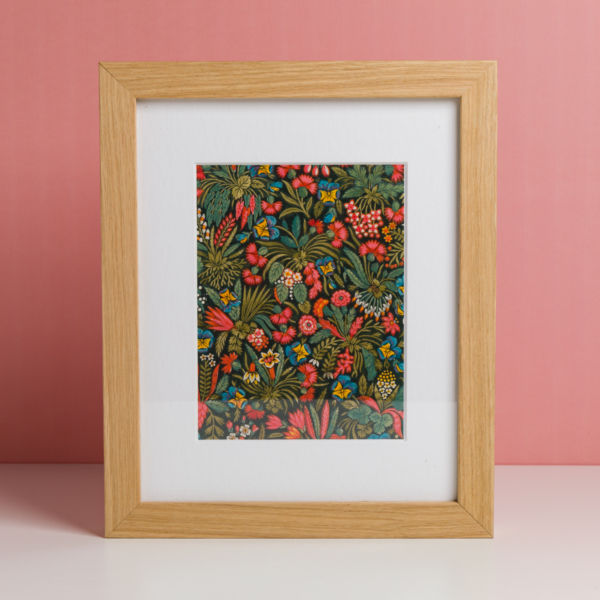 L'EMPREINTE SOUS PASSE-PARTOUT "BONNES HERBES - BLEUETS" est une œuvre d'art encadrée avec des fleurs rouges, jaunes et bleues et des feuilles vertes sur un fond sombre, placée dans un cadre en bois clair et présentée sur une surface blanche avec un arrière-plan rose. la marque Toile de Jouy
