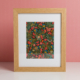 L'EMPREINTE SOUS PASSE-PARTOUT "BONNES HERBES - BLEUETS" est une œuvre d'art encadrée avec des fleurs rouges, jaunes et bleues et des feuilles vertes sur un fond sombre, placée dans un cadre en bois clair et présentée sur une surface blanche avec un arrière-plan rose. la marque Toile de Jouy