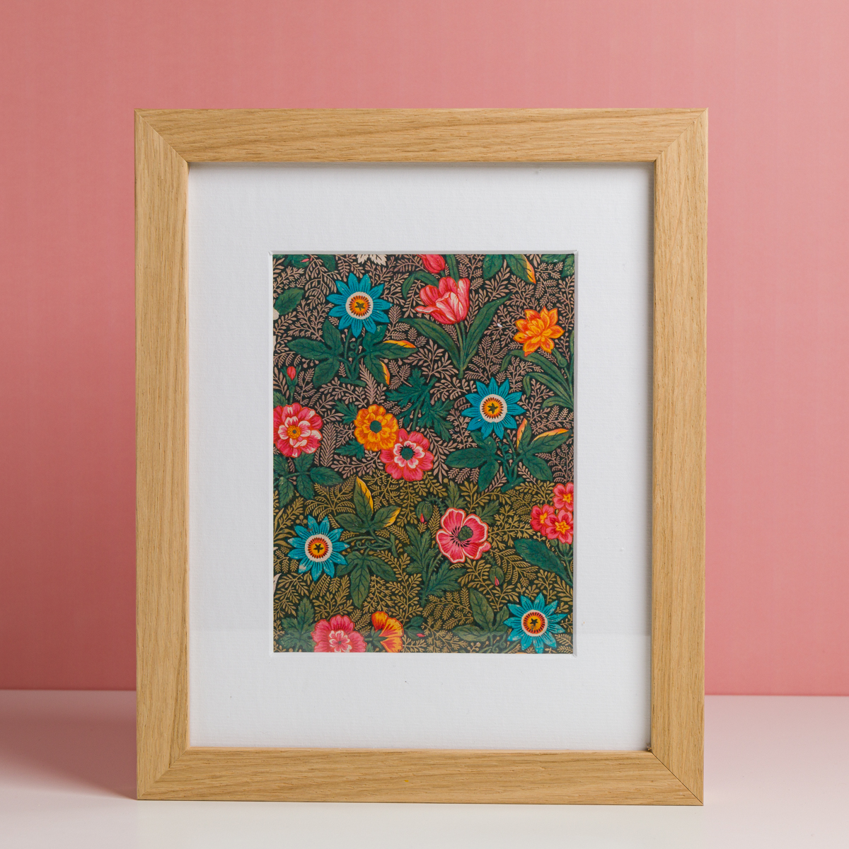L'EMPREINTE SOUS PASSE-PARTOUT "BONNES HERBES - BLEUETS" présente une illustration florale vibrante avec des feuilles vertes et des fleurs colorées sur un fond rose, présentée dans un cadre en bois sur une surface blanche. la marque Toile de Jouy