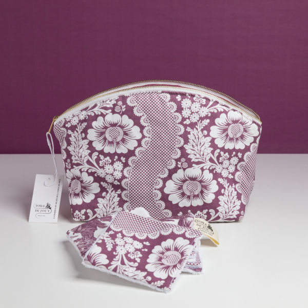 La pochette XCOMM_TROUSSE ARRONDIE FLEURS AMARANTE, une pochette à fermeture éclair à motif floral violet et blanc, accompagnée de disques démaquillants réutilisables assortis et d’une étiquette de marque, est présentée sur une surface blanche devant un fond violet. la marque Toile de Jouy