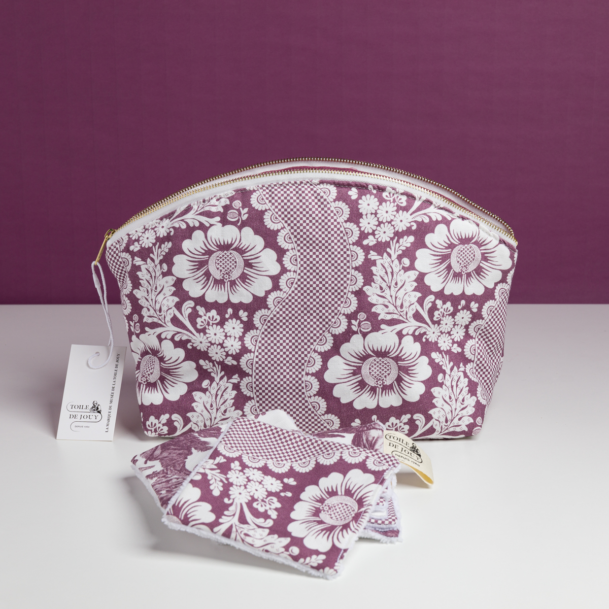 La pochette XCOMM_TROUSSE ARRONDIE FLEURS AMARANTE, une pochette à fermeture éclair à motif floral violet et blanc, accompagnée de disques démaquillants réutilisables assortis et d’une étiquette de marque, est présentée sur une surface blanche devant un fond violet. la marque Toile de Jouy