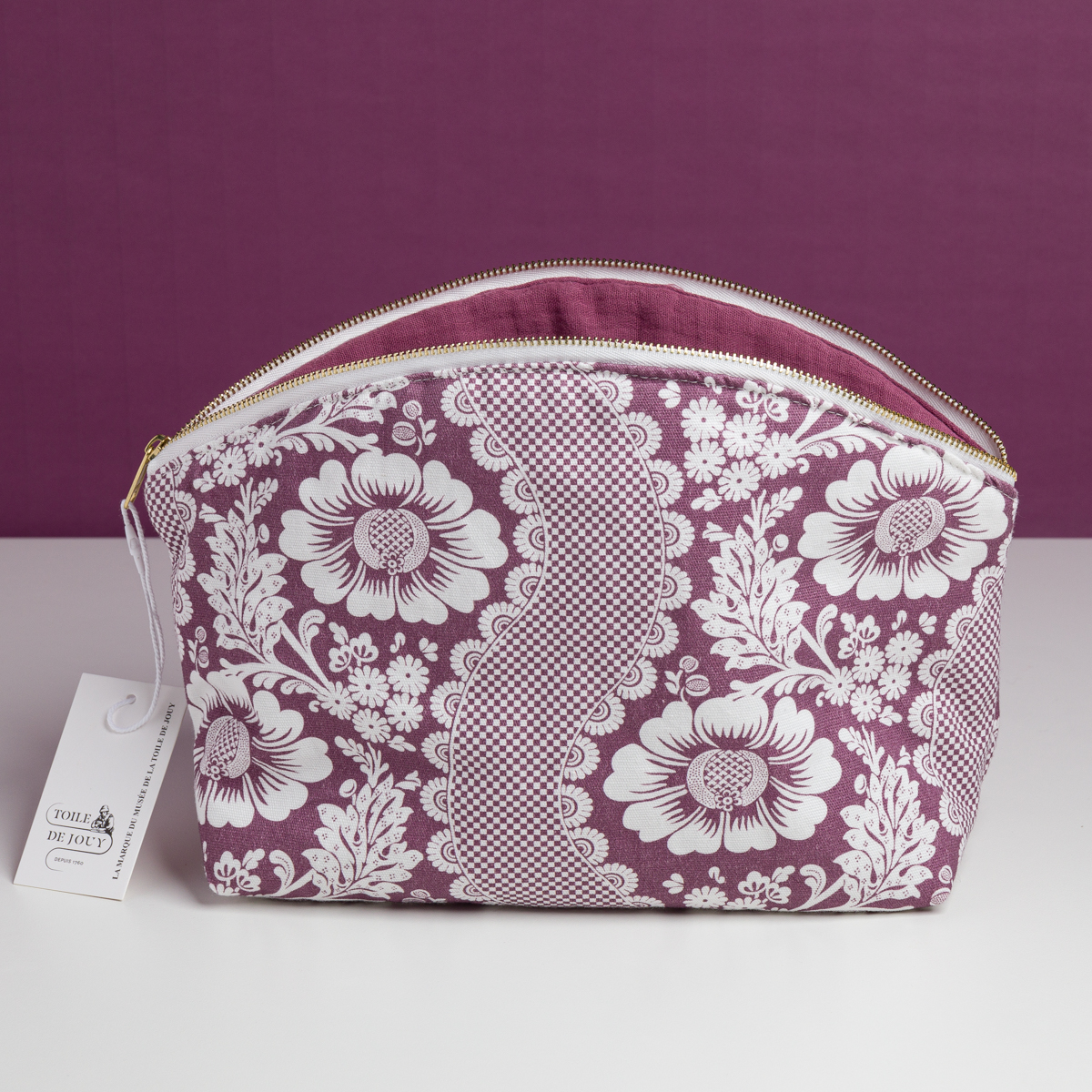 La pochette XCOMM_TROUSSE ARRONDIE FLEURS AMARANTE, une pochette à fermeture éclair ornée d’un motif floral violet et blanc et dotée d’une étiquette sur la tirette gauche de la fermeture éclair, repose sur une surface blanche devant un fond violet assorti. la marque Toile de Jouy