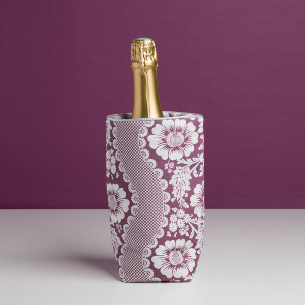 Une bouteille de champagne dans un manchon XCOMM_SET DE TABLE /2 FLEURS STYLISEES SAUGE à motif floral, sur un fond violet assorti, sur une surface blanche. la marque Toile de Jouy