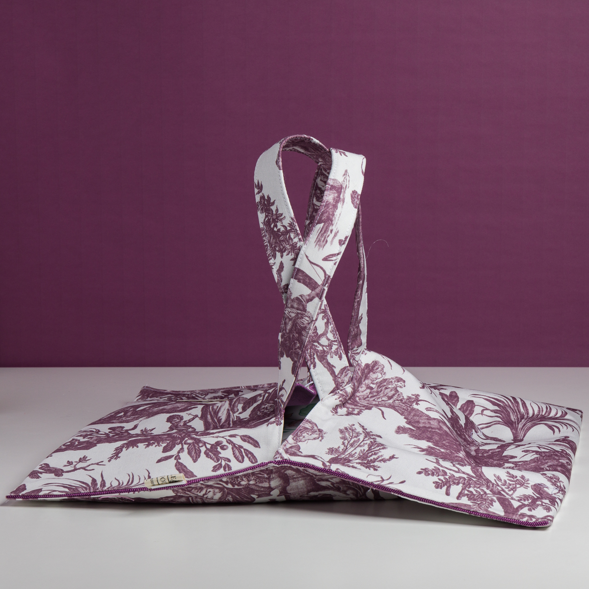 Le XCOMM_SET DE TABLE /2 FLEURS STYLISEES SAUGE, qui présente des motifs floraux stylisés de sauge sur fond blanc, est présenté partiellement déplié sur une surface blanche avec un fond coordonné à motifs violets. la marque Toile de Jouy