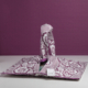 Le XCOMM_SET DE TABLE /2 FLEURS STYLISEES SAUGE, un sac en tissu à motifs floraux avec des poignées et une étiquette blanche, repose sur une surface blanche sur un fond violet coordonné. la marque Toile de Jouy
