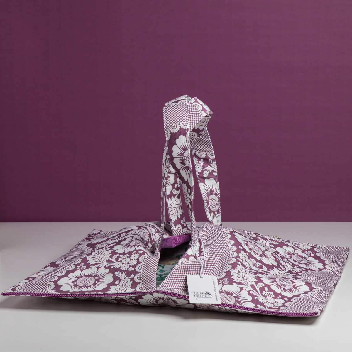 Le XCOMM_SET DE TABLE /2 FLEURS STYLISEES SAUGE, un sac en tissu à motifs floraux avec des poignées et une étiquette blanche, repose sur une surface blanche sur un fond violet coordonné. la marque Toile de Jouy