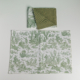 Le XCOMM_SET DE TABLE /2 OFFRANDE SAUGE comprend deux sets de table en tissu avec des scènes pastorales vertes sur fond texturé blanc et vert uni ; l'un est plié pour présenter les deux faces, l'autre est posé à plat sur une surface blanche. la marque Toile de Jouy
