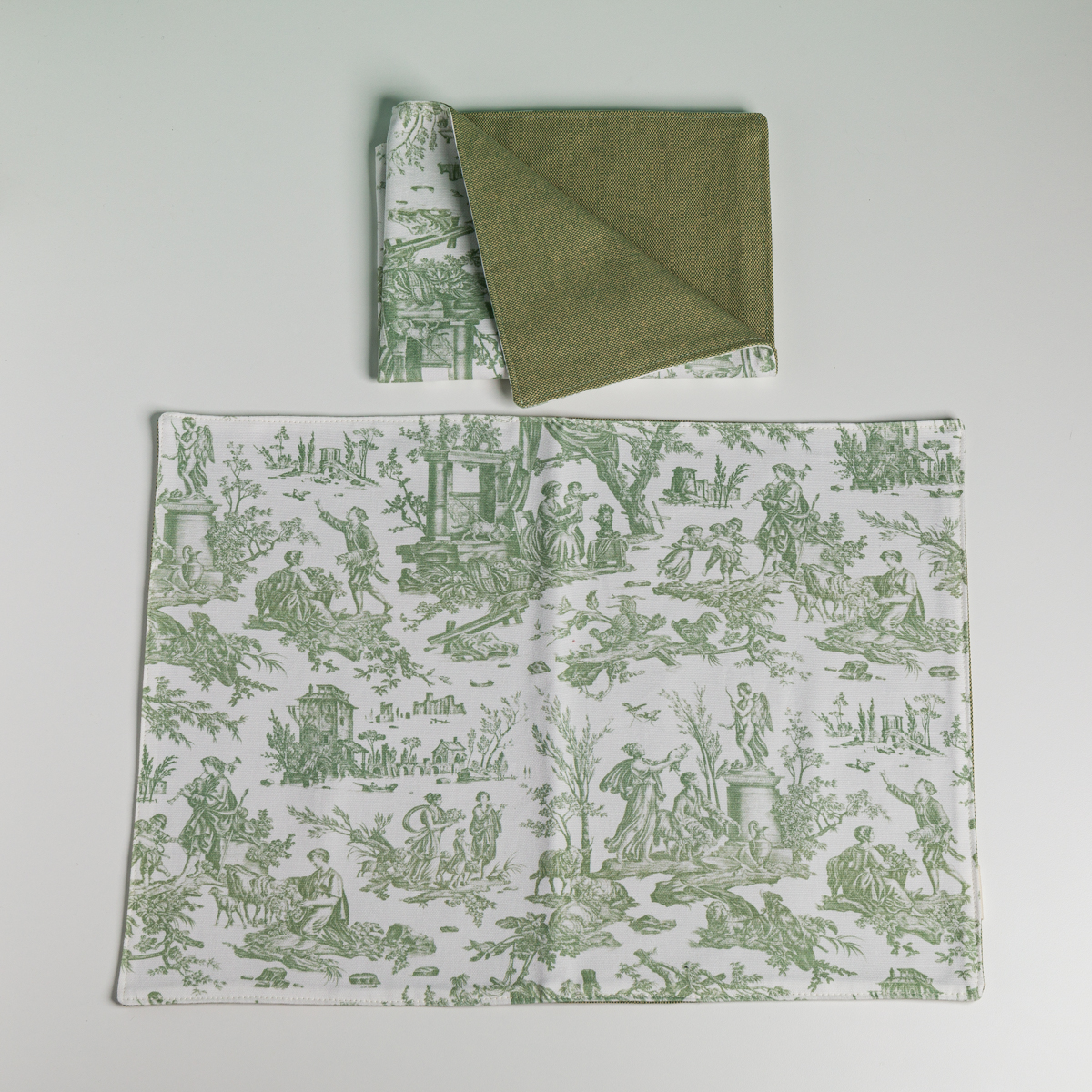 Le XCOMM_SET DE TABLE /2 OFFRANDE SAUGE comprend deux sets de table en tissu avec des scènes pastorales vertes sur fond texturé blanc et vert uni ; l'un est plié pour présenter les deux faces, l'autre est posé à plat sur une surface blanche. la marque Toile de Jouy