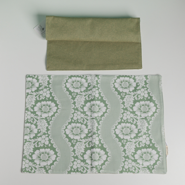Le XCOMM_SET DE TABLE /2 FLEURS STYLISEES SAUGE comprend une pochette en tissu vert uni posée sur un set de table assorti vert sauge et blanc avec un motif de dentelle et de fleurs, le tout sur fond blanc. la marque Toile de Jouy