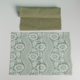 Le XCOMM_SET DE TABLE /2 FLEURS STYLISEES SAUGE comprend une pochette en tissu vert uni posée sur un set de table assorti vert sauge et blanc avec un motif de dentelle et de fleurs, le tout sur fond blanc. la marque Toile de Jouy