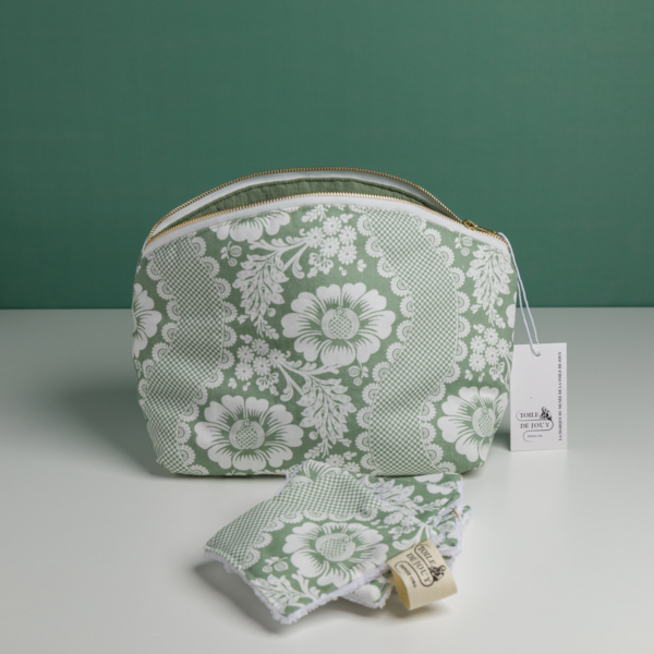 La pochette XCOMM_TROUSSE ARRONDIE FLEURS AMARANTE, une pochette à motif floral vert et blanc dotée d'une fermeture à glissière et d'une étiquette portant le logo de la marque, est posée à la verticale sur une surface blanche. Des lingettes en tissu assorties sont disposées devant, sur un fond vert uni. la marque Toile de Jouy