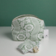 La pochette XCOMM_TROUSSE ARRONDIE FLEURS AMARANTE, une pochette à motif floral vert et blanc dotée d'une fermeture à glissière et d'une étiquette portant le logo de la marque, est posée à la verticale sur une surface blanche. Des lingettes en tissu assorties sont disposées devant, sur un fond vert uni. la marque Toile de Jouy