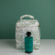 Le parfum XCOMM_VANITY OFFRANDE SAUGE se présente dans un flacon en verre bleu sarcelle muni d'un bouchon noir, posé devant une trousse de toilette blanche à fermeture éclair ornée d'un imprimé champêtre vert, le tout sur un fond vert uni. la marque Toile de Jouy