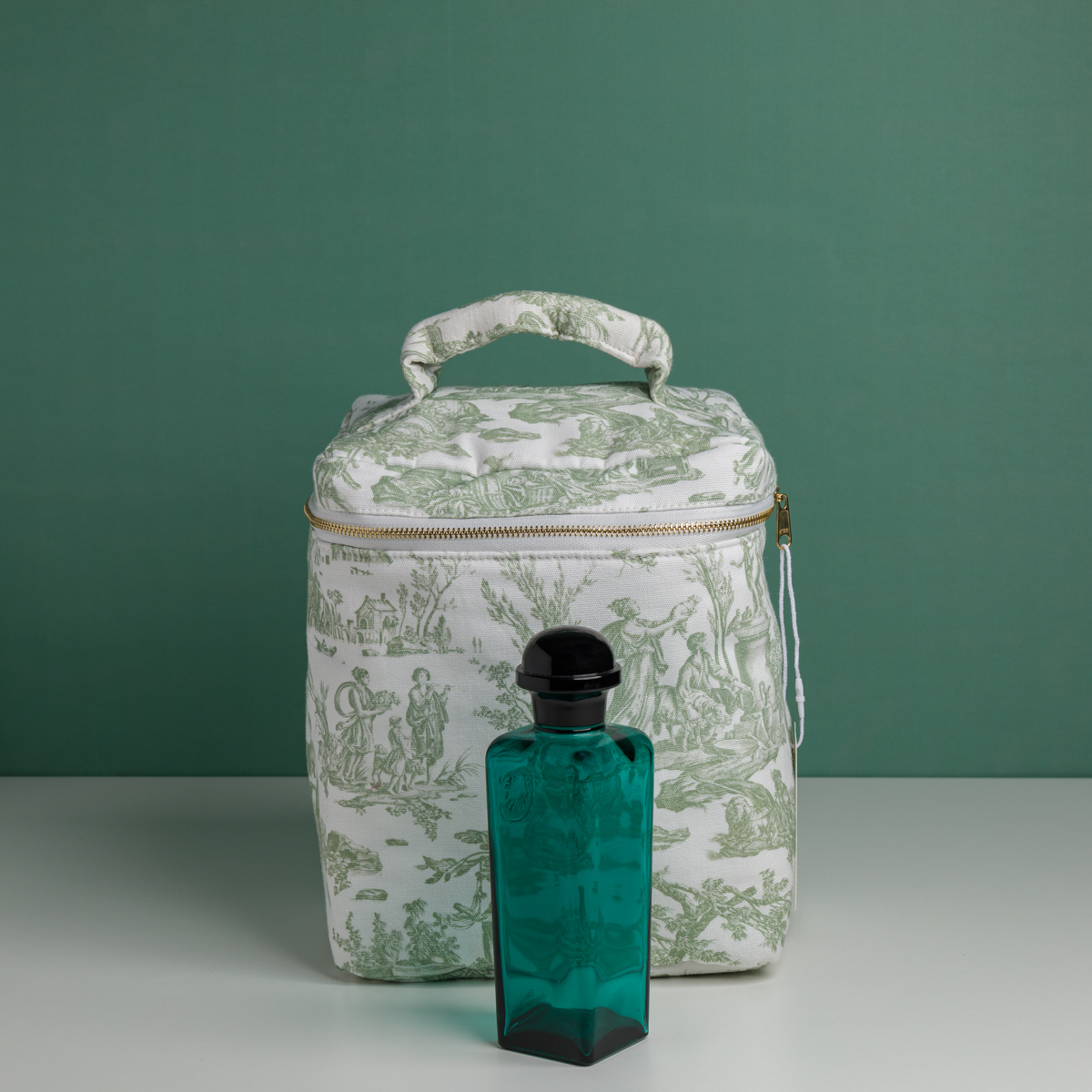 Le parfum XCOMM_VANITY OFFRANDE SAUGE se présente dans un flacon en verre bleu sarcelle muni d'un bouchon noir, posé devant une trousse de toilette blanche à fermeture éclair ornée d'un imprimé champêtre vert, le tout sur un fond vert uni. la marque Toile de Jouy