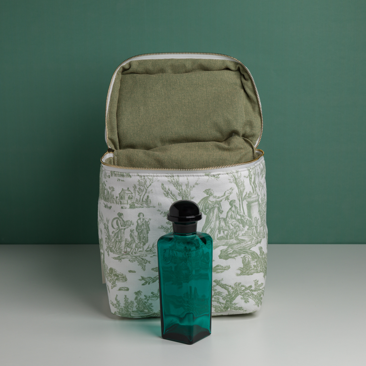 Le parfum XCOMM_VANITY OFFRANDE SAUGE se présente dans un flacon en verre bleu sarcelle muni d'un bouchon noir, posé devant une pochette en tissu à motifs, dont l'intérieur est vert et la fermeture éclair dorée, le tout sur fond vert. la marque Toile de Jouy