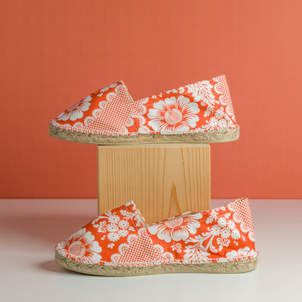 Le modèle XCOMM_ESPADRILLE FLEURS MANDARINE P38, orné de motifs floraux blancs, est présenté avec une chaussure posée sur un bloc en bois et l'autre sur une surface blanche, le tout sur fond orange. la marque Toile de Jouy
