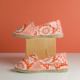 Le modèle XCOMM_ESPADRILLE FLEURS MANDARINE P38, orné de motifs floraux blancs, est présenté avec une chaussure posée sur un bloc en bois et l'autre sur une surface blanche, le tout sur fond orange. la marque Toile de Jouy