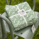 Le XCOMM_NAPPE FORAL SAUGE 180X180, un tissu vert et blanc à motifs floraux, est noué avec un ruban blanc et présenté à l'extérieur sur une chaise métallique grise posée sur de l'herbe. la marque Toile de Jouy