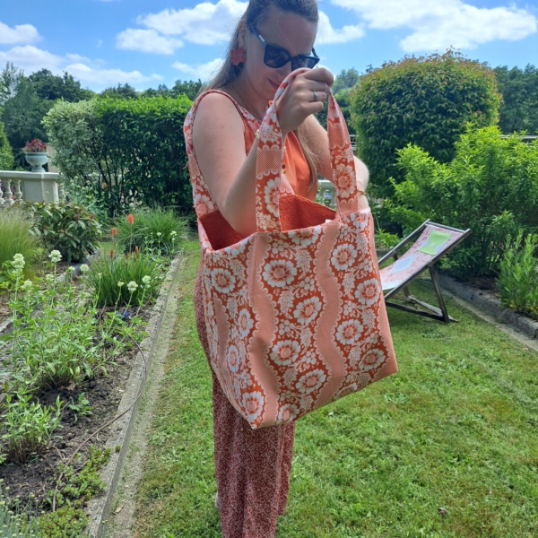 Une femme vêtue d'un ensemble rose à motifs se tient debout dans un jardin, tenant à la main le sac fourre-tout « XCOMM_SAC DE PLAGE ENDUIT-EPONGE FLEURS MANDARINE » ; elle regarde à l'intérieur du sac, avec en arrière-plan des plantes vertes, une chaise longue et un ciel partiellement nuageux. la marque Toile de Jouy
