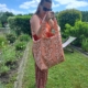 Une femme vêtue d'un ensemble rose à motifs se tient debout dans un jardin, tenant à la main le sac fourre-tout « XCOMM_SAC DE PLAGE ENDUIT-EPONGE FLEURS MANDARINE » ; elle regarde à l'intérieur du sac, avec en arrière-plan des plantes vertes, une chaise longue et un ciel partiellement nuageux. la marque Toile de Jouy