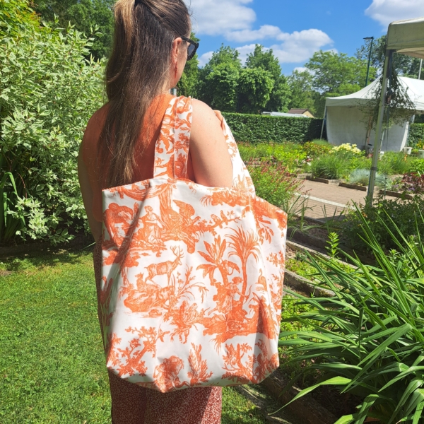 Une femme aux longs cheveux châtains, portant des lunettes de soleil, se tient dans un jardin ensoleillé, tenant dans ses mains le XCOMM_SAC DE PLAGE ENDUIT-EPONGE FLEURS MANDARINE — un grand sac fourre-tout blanc orné d’un motif floral orange. Derrière elle, on aperçoit une végétation luxuriante et une tente blanche. la marque Toile de Jouy