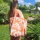 Une femme aux longs cheveux châtains, portant des lunettes de soleil, se tient dans un jardin ensoleillé, tenant dans ses mains le XCOMM_SAC DE PLAGE ENDUIT-EPONGE FLEURS MANDARINE — un grand sac fourre-tout blanc orné d’un motif floral orange. Derrière elle, on aperçoit une végétation luxuriante et une tente blanche. la marque Toile de Jouy