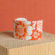 Le modèle XCOMM_ESPADRILLE FLEURS MANDARINE P38 est un bracelet large à revers blanc, orné de motifs floraux et botaniques orange vif, présentés sur un bloc de bois clair avec un fond orange. la marque Toile de Jouy