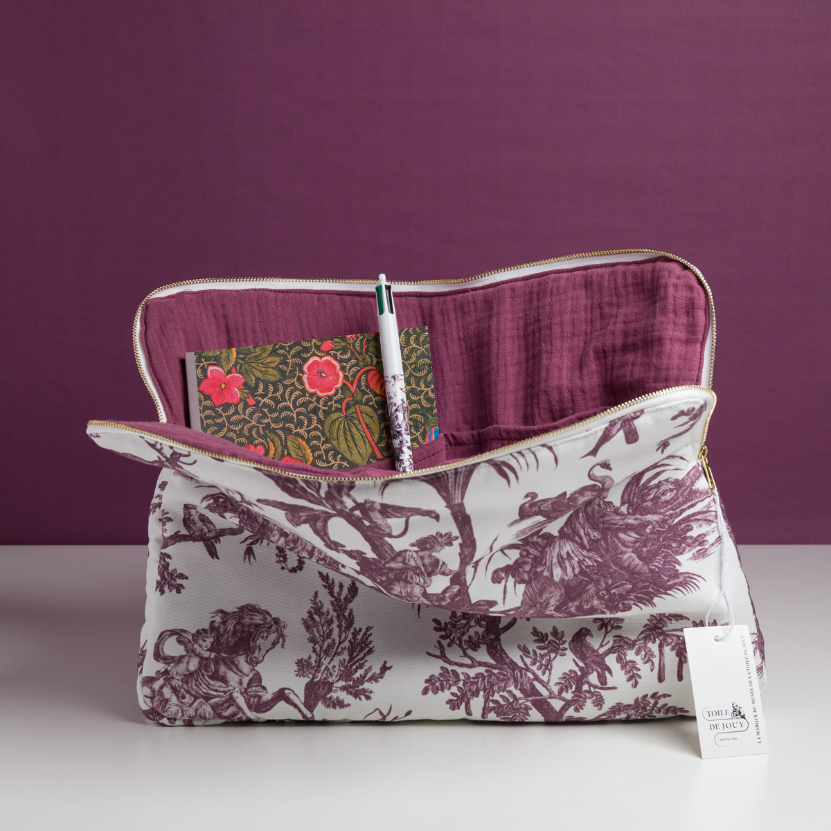 Le XCOMM_HOUSSE ORDI 4 APRTIES AMARANTE, qui présente un motif mythologique violet et blanc, est ouvert sur une surface blanche, avec à l'intérieur un carnet de notes à fleurs et un stylo. Un fond violet uni complète la scène. la marque Toile de Jouy
