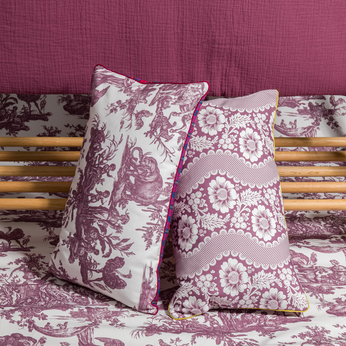 Les oreillers décoratifs XCOMM_COUSSIN 30X50 FLEURS SAUGE reposent sur une tête de lit en bois au sommet d'un lit avec une literie et une couverture à motifs violets et blancs assortis. la marque Toile de Jouy