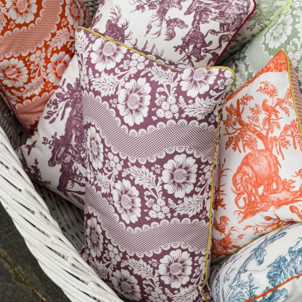 Le coussin décoratif XCOMM_COUSSIN 30X50 FLEURS AMARANTE, aux motifs floraux complexes, est disposé dans un panier tressé blanc. la marque Toile de Jouy