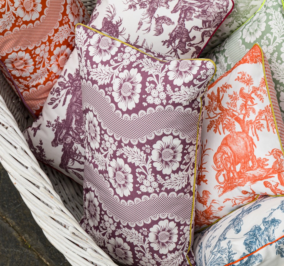 Le coussin décoratif XCOMM_COUSSIN 30X50 FLEURS AMARANTE, aux motifs floraux complexes, est disposé dans un panier tressé blanc. la marque Toile de Jouy