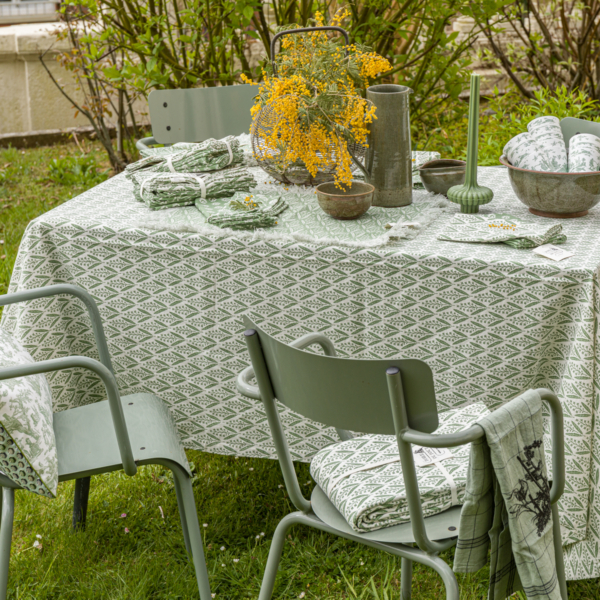 Une table de jardin présente la nappe verte et blanche XCOMM_NAPPE FORAL SAUGE 180X180, des serviettes de table assorties, des chaises vertes, des fleurs jaunes dans un vase, des bols, un chandelier vert et de la verdure en arrière-plan. la marque Toile de Jouy